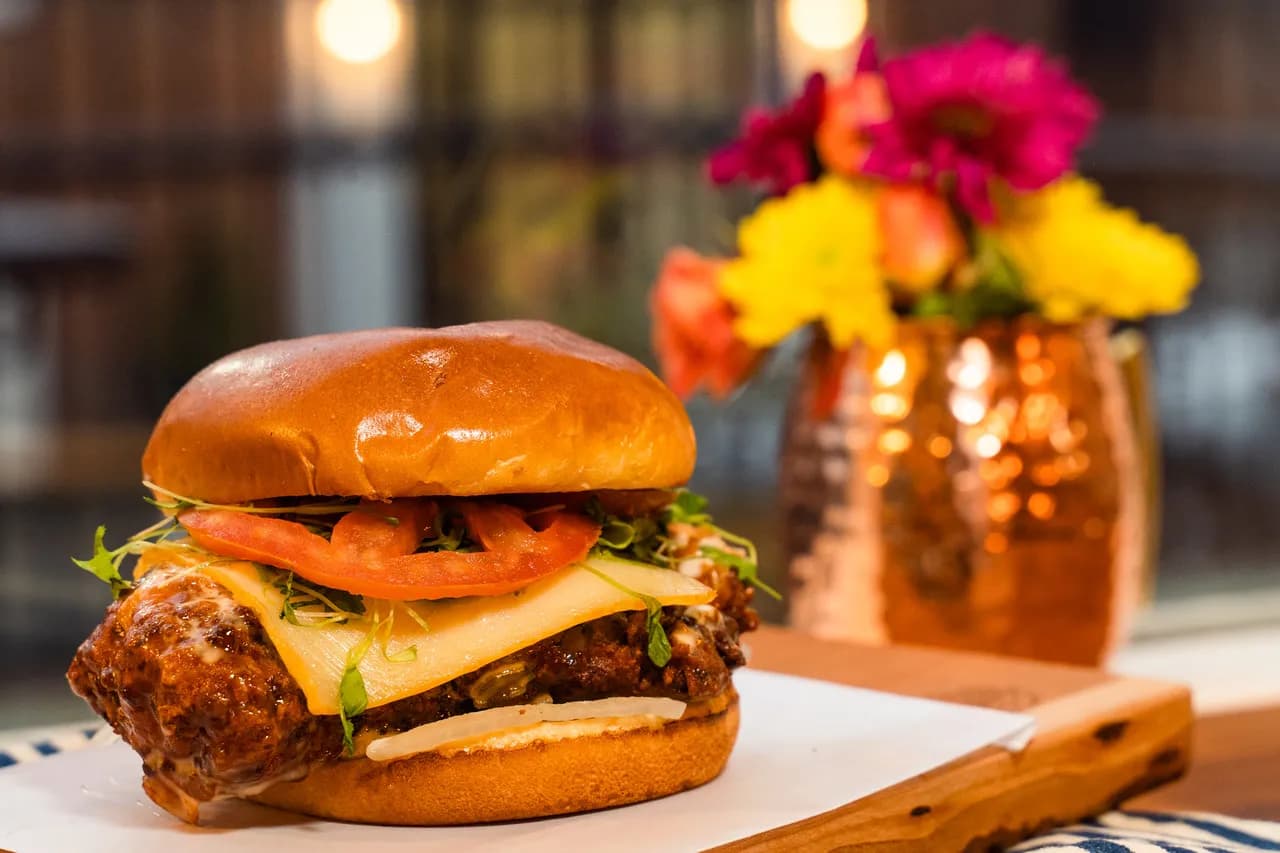This Chicken Flew Southwest — fried marinated chicken breast with Muenster, tomato, micro cilantro, and spicy serrano pepper honey ranch on a brioche bun, photographed warm with a copper mule and floral arrangement at The Burgerly.
