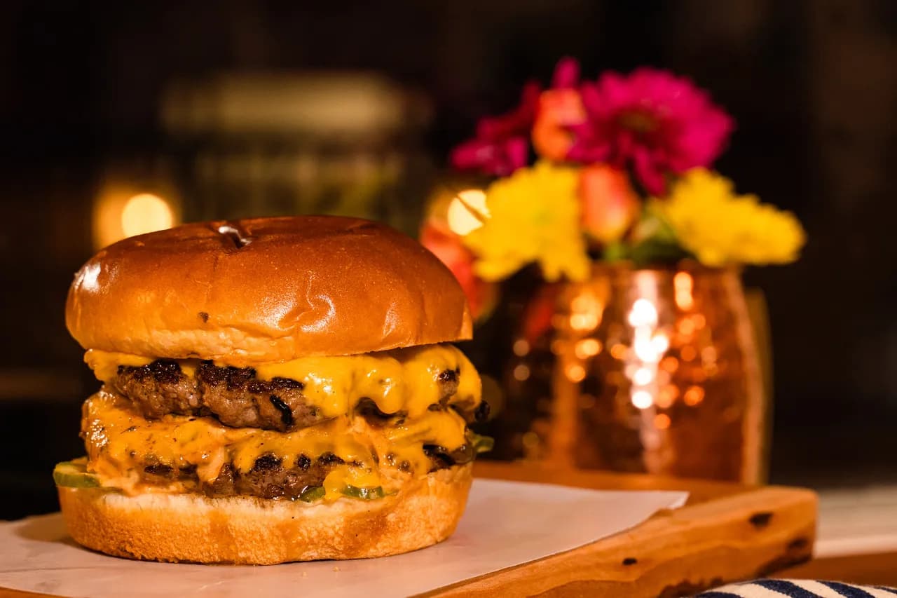 The Burgerly — cheeseburger on a brioche bun with savory onions and house pickles, plated warm on a live-edge wood board.