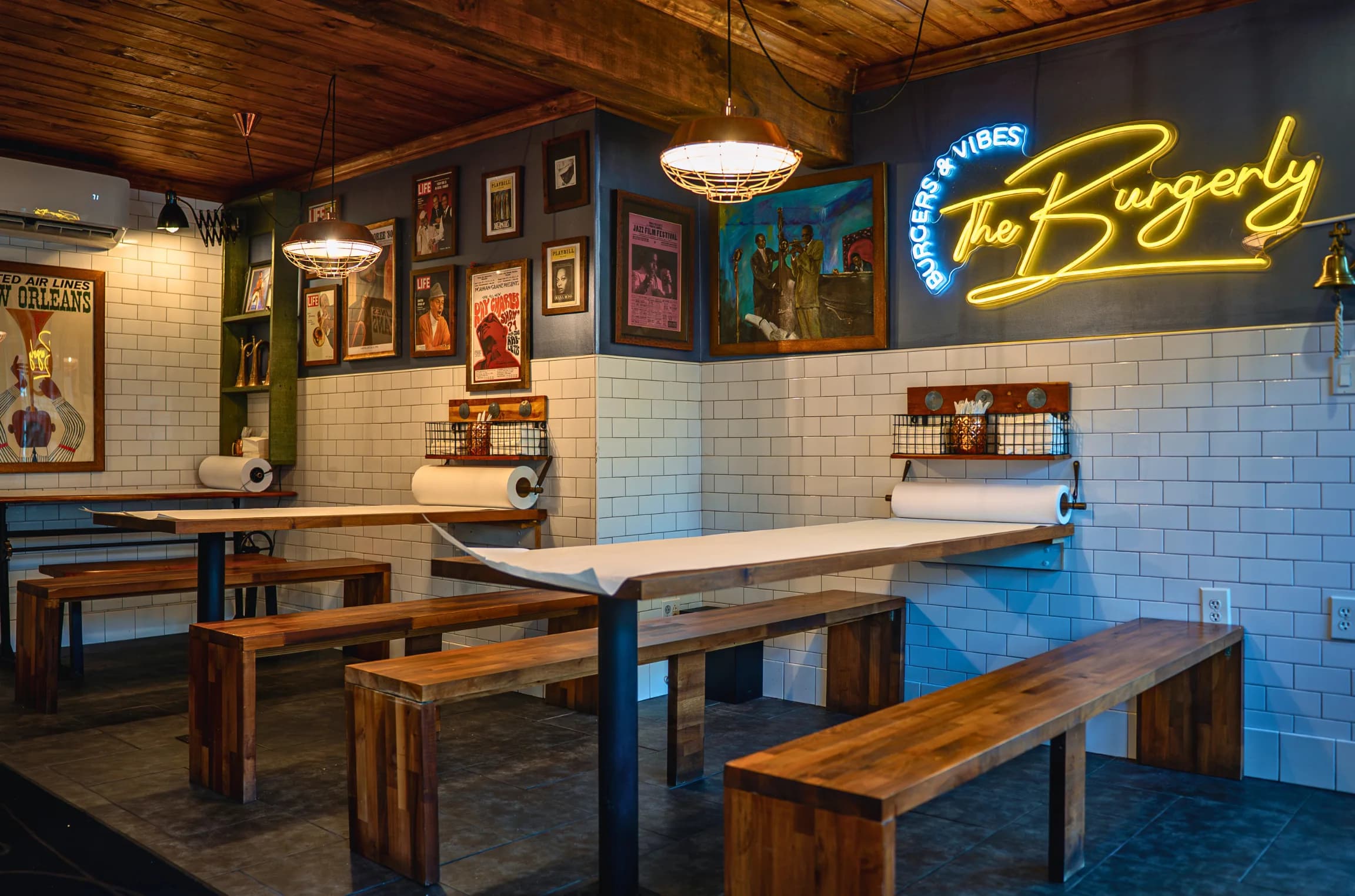 The Burgerly's framed wall — jazz posters, vintage magazine covers, the Picasso-style painting, and the yellow neon Burgerly sign over wood ceiling and white subway tile.