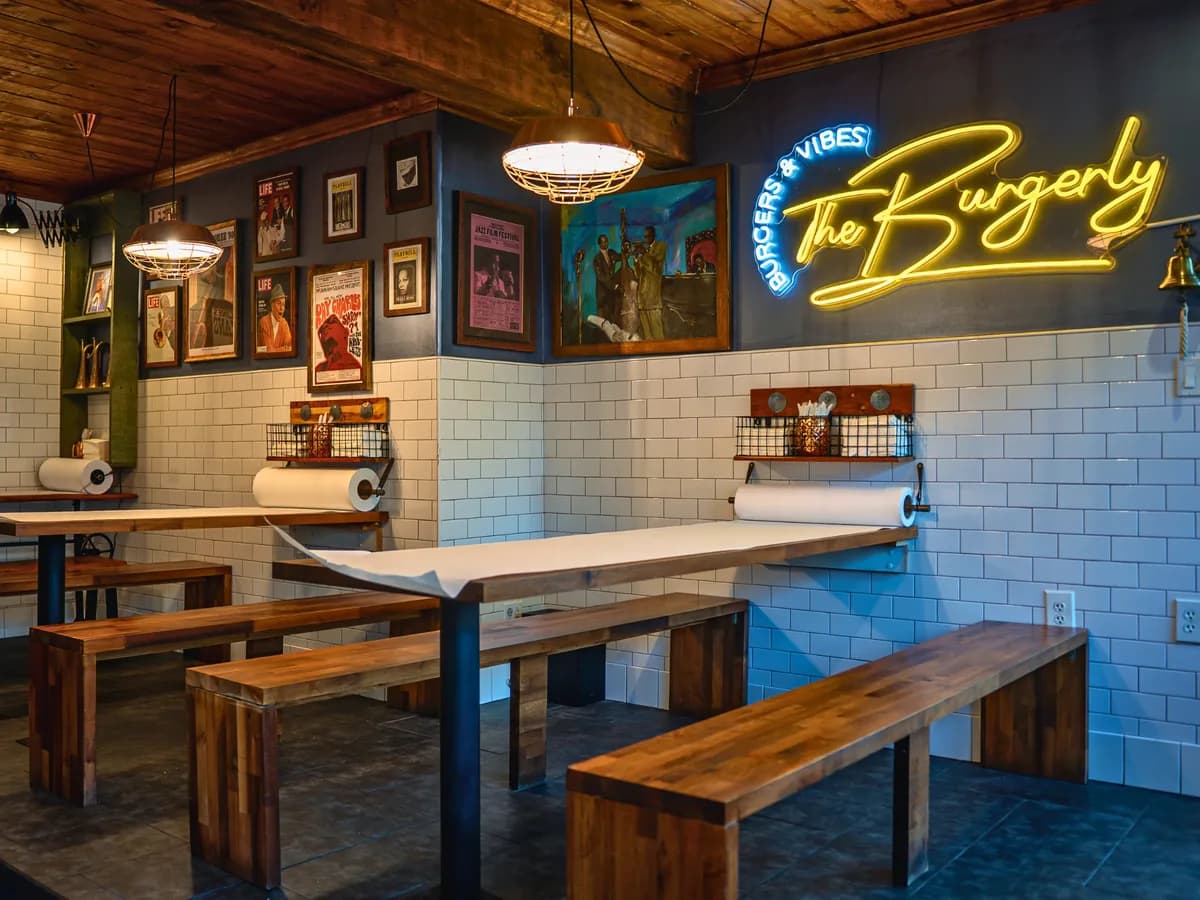 Inside The Burgerly — long communal tables with parchment rolls and benches, under the jazz wall and the yellow neon Burgerly sign.