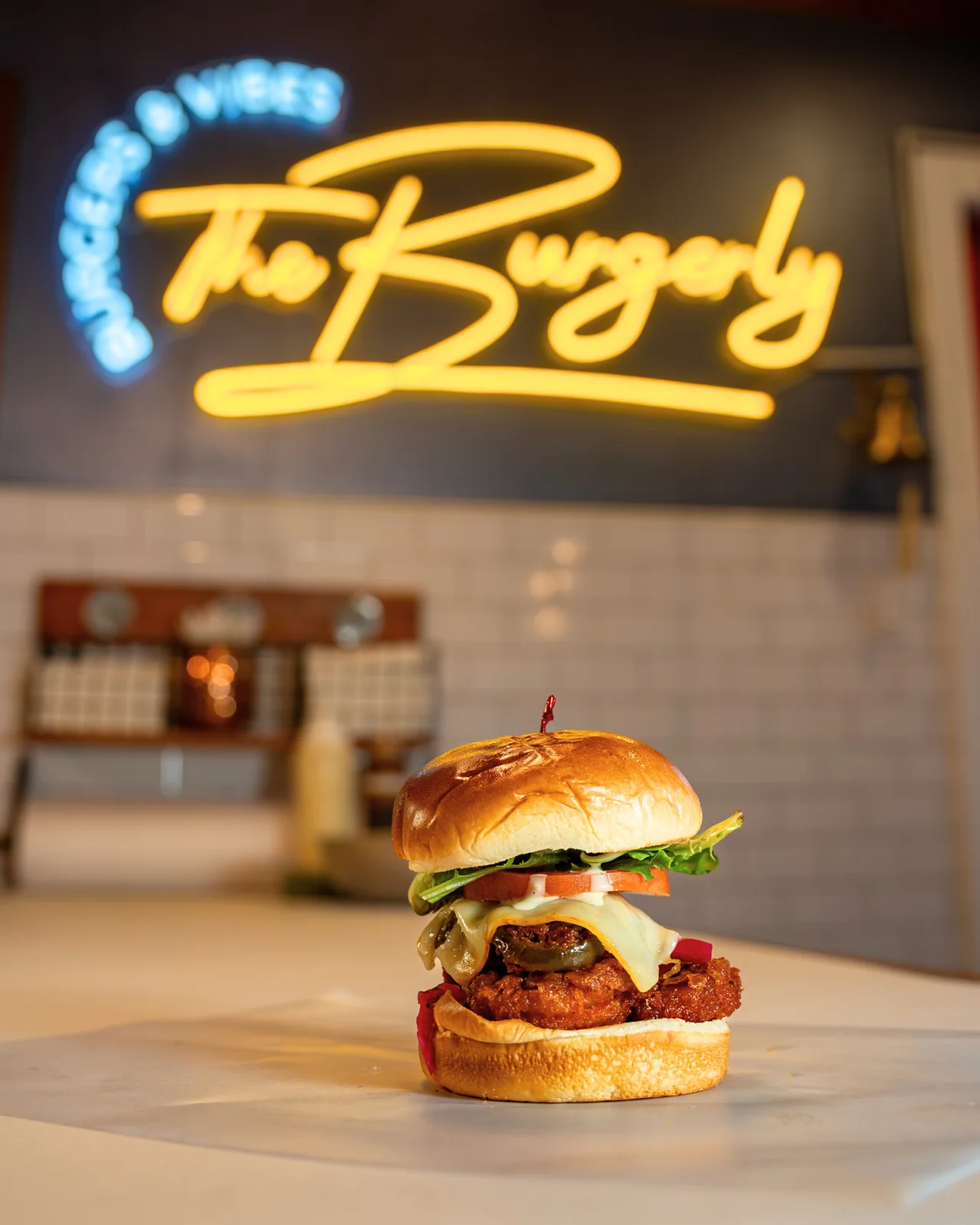 Chicken Flew Southwest — a fried marinated chicken sandwich with Muenster, pickled onions, and serrano honey ranch on a brioche bun, plated under the yellow neon Burgerly sign.