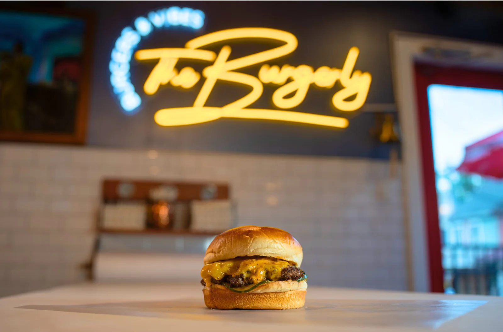 The Burgerly — a classic brioche cheeseburger on butcher block, the yellow neon Burgerly sign glowing on the back wall.