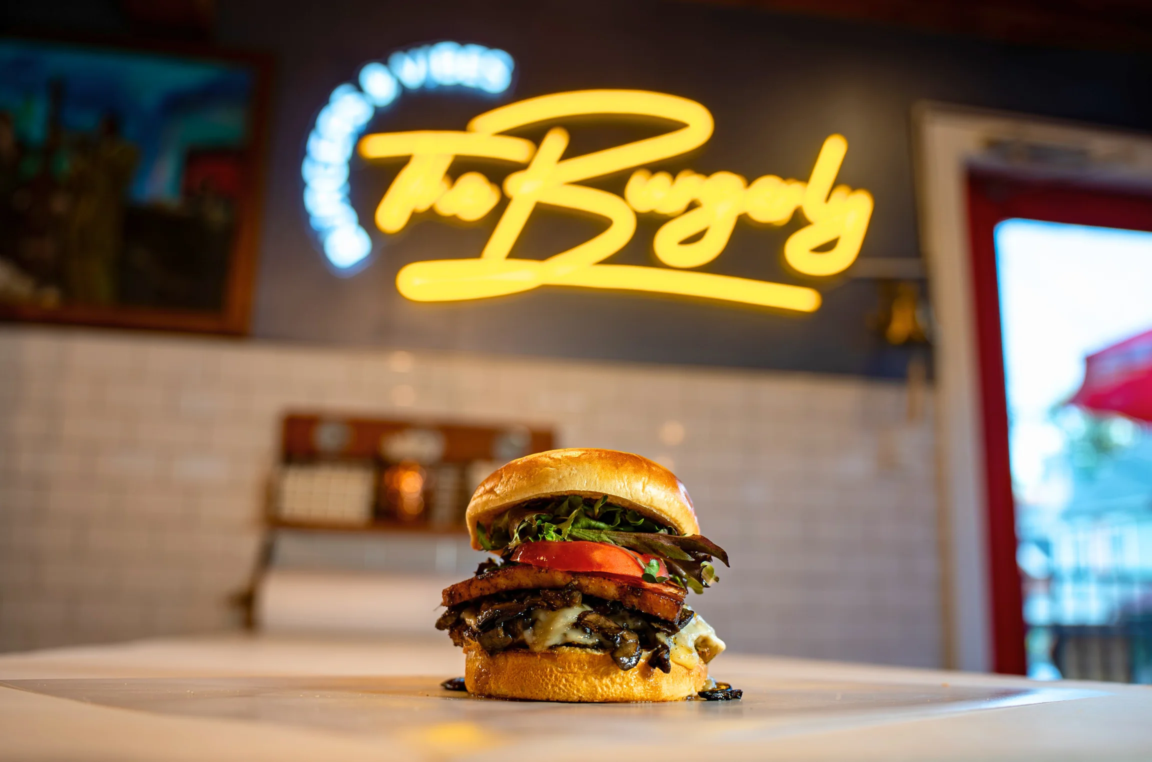The Wonderful — a chef-driven cheeseburger with house Canadian bacon, savory onions, Havarti, and greens on a brioche bun, photographed inside The Burgerly with the yellow neon "The Burgerly — Burgers & Vibes" sign glowing on the back wall.
