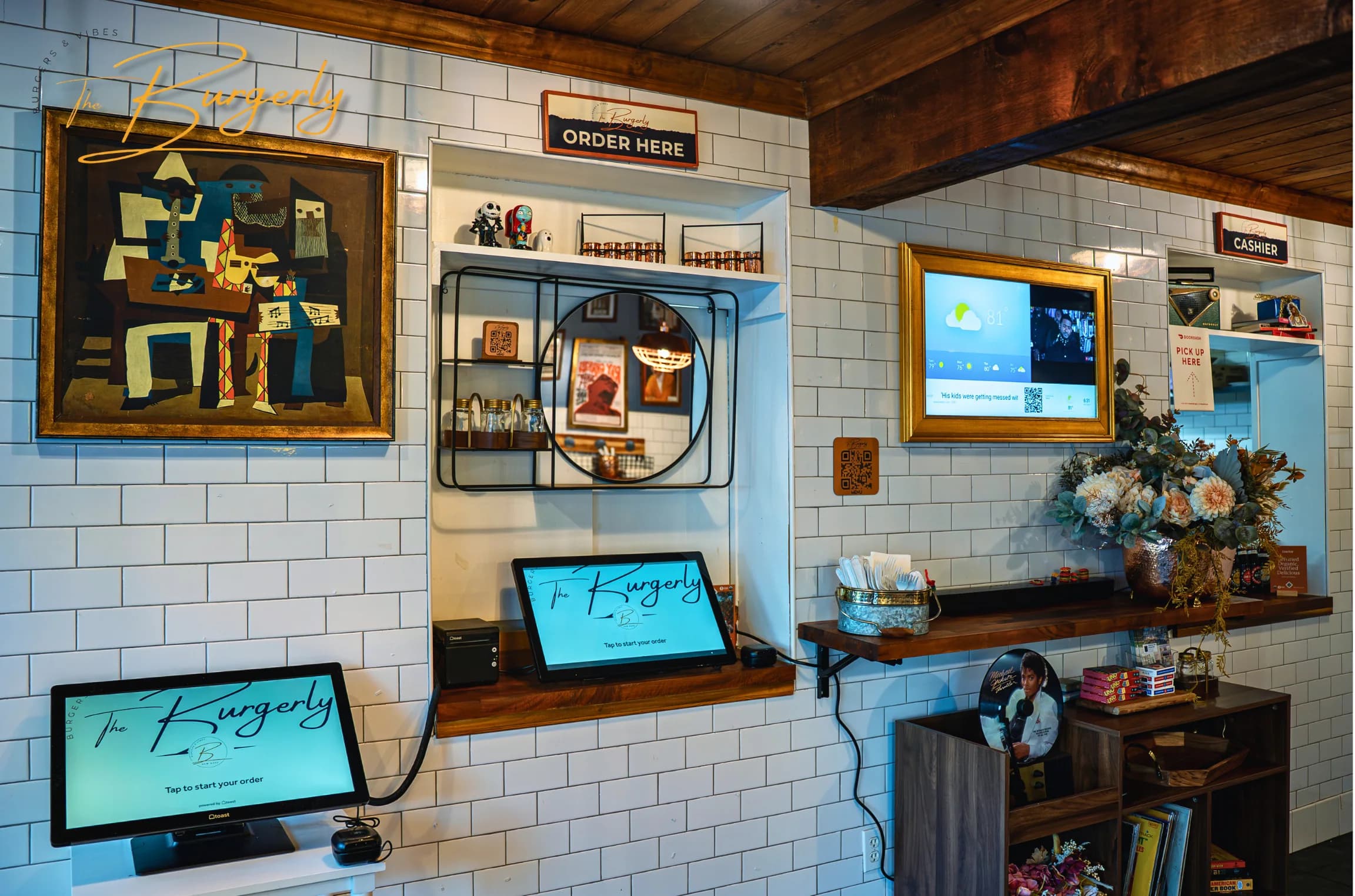 Detail of the curated wall at The Burgerly — the Picasso-style painting, a small mirror, framed art, and the order screens reading 'The Burgerly,' grouped on white subway tile.