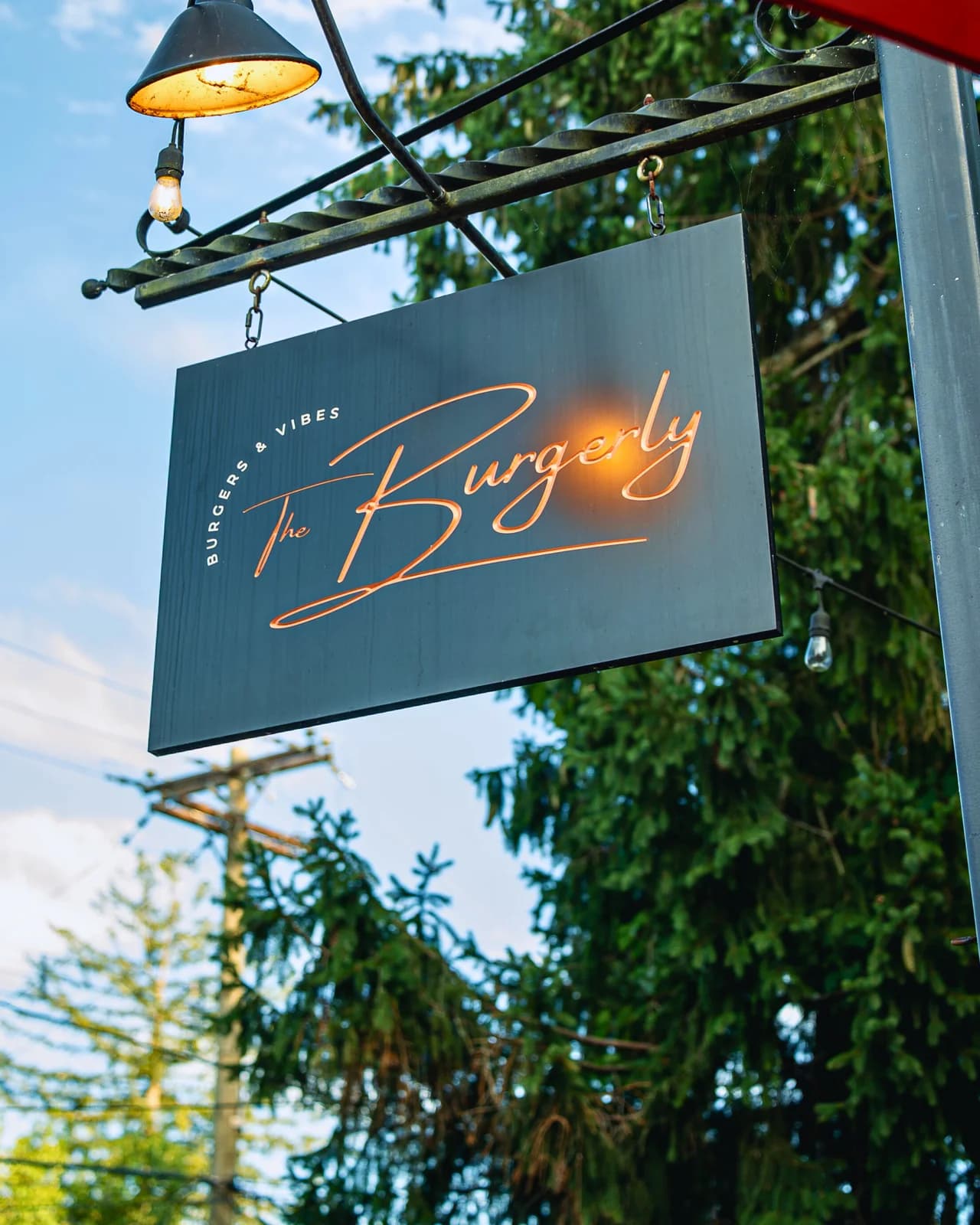 The Burgerly's hanging copper-script wordmark sign — 'Burgers & Vibes' above 'The Burgerly' — glowing under a warm Edison bulb at golden hour, framed by evergreen trees and the blue sky above S. Main Street.