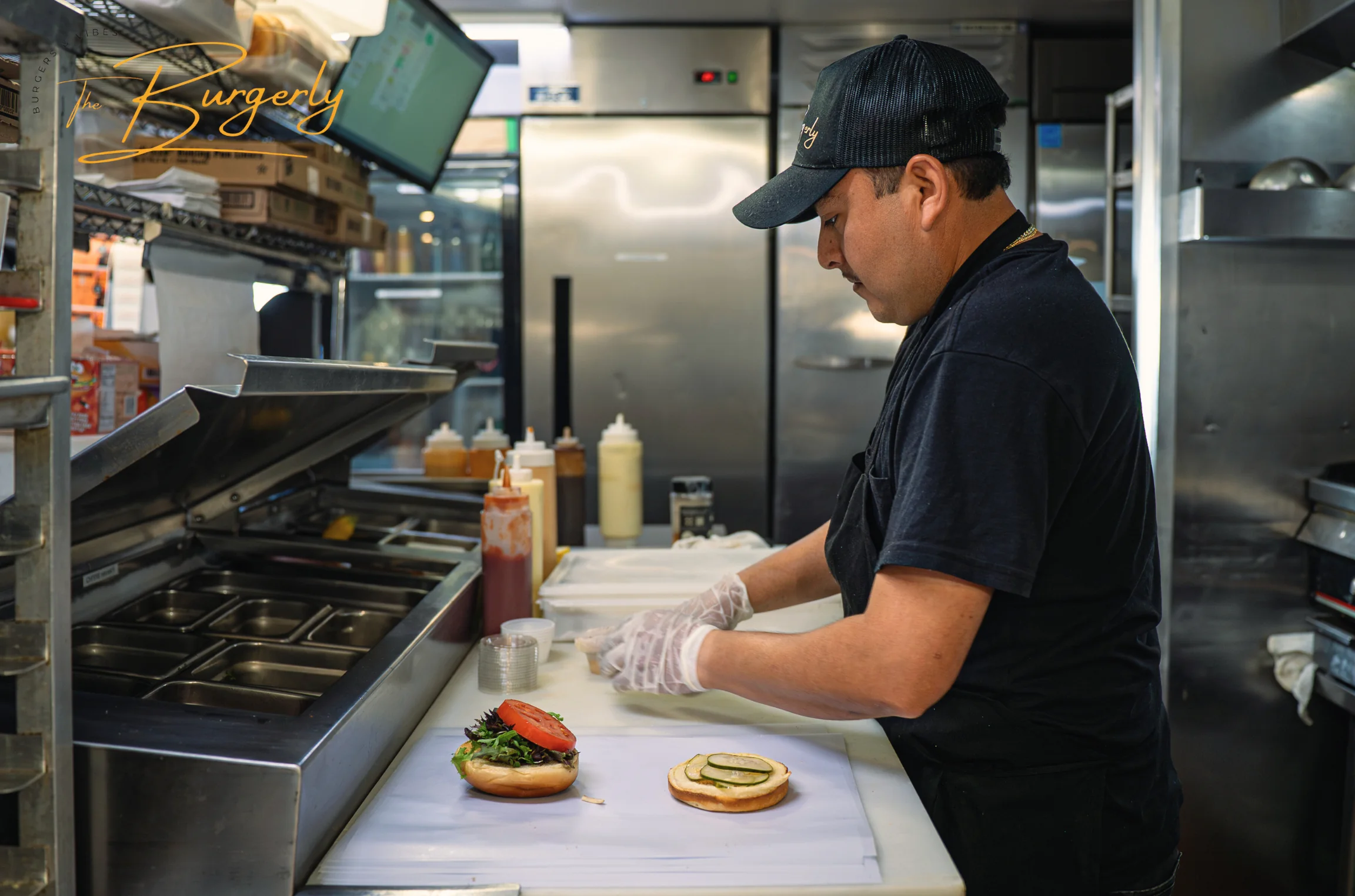 Chef Mark plating at the kitchen pass — gloved hands building a brioche-bun cheeseburger with greens and tomato over stainless prep.
