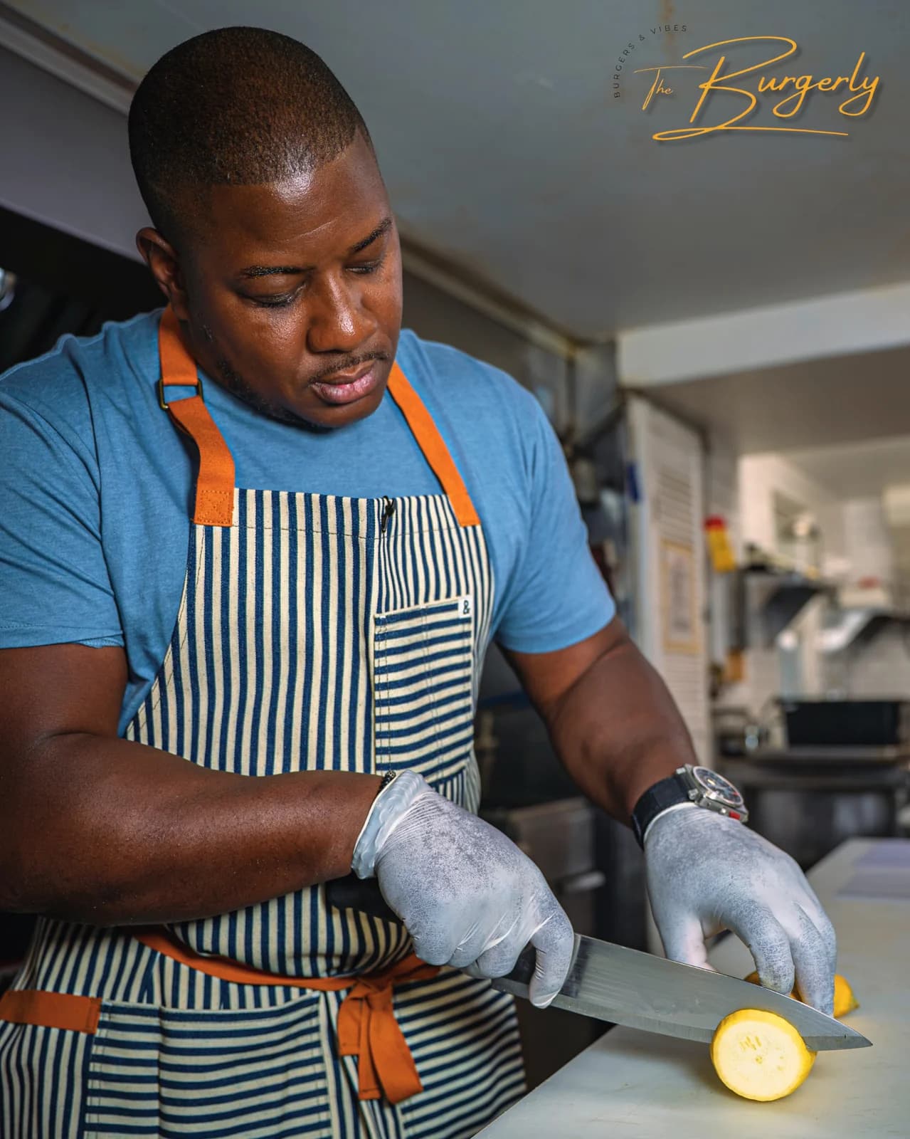Chef Mark McLean at work in The Burgerly kitchen — blue tee, blue-and-orange striped chef apron, gloved hands, knife on a lemon, focused.